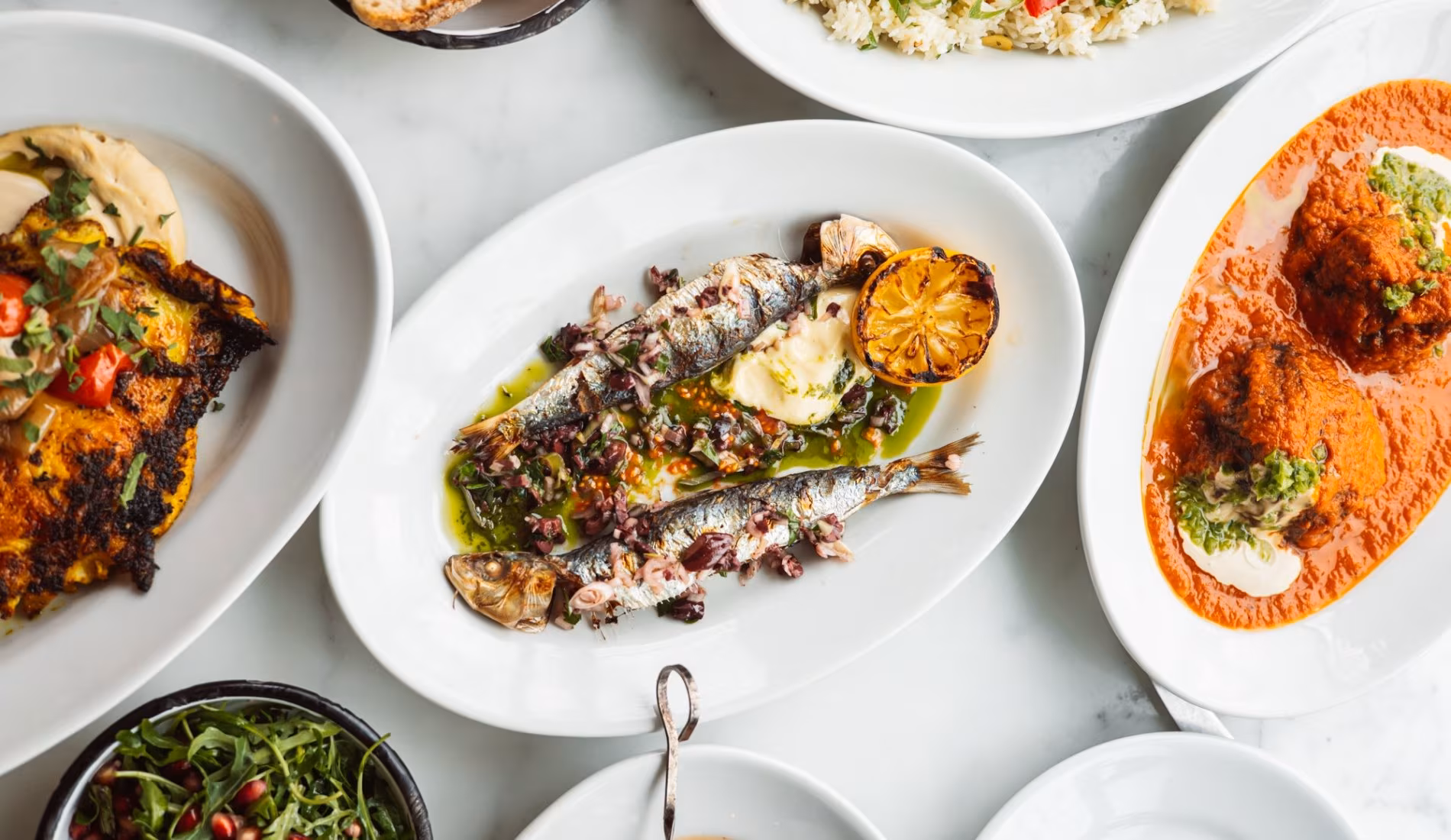 Plates of grilled vegetables, greens and dips on a white table