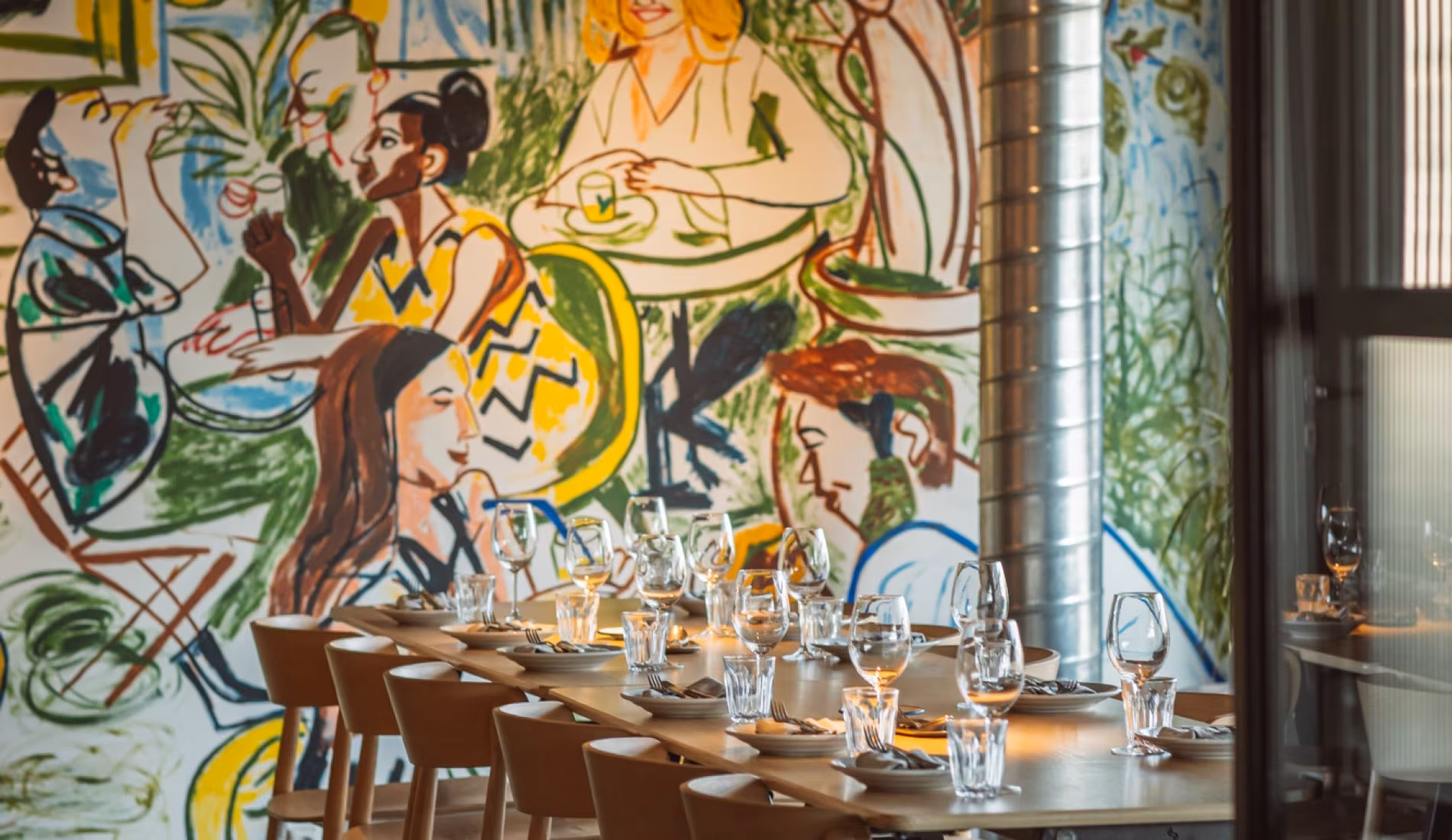 Long communal table set with glassware in front of a colourful mural