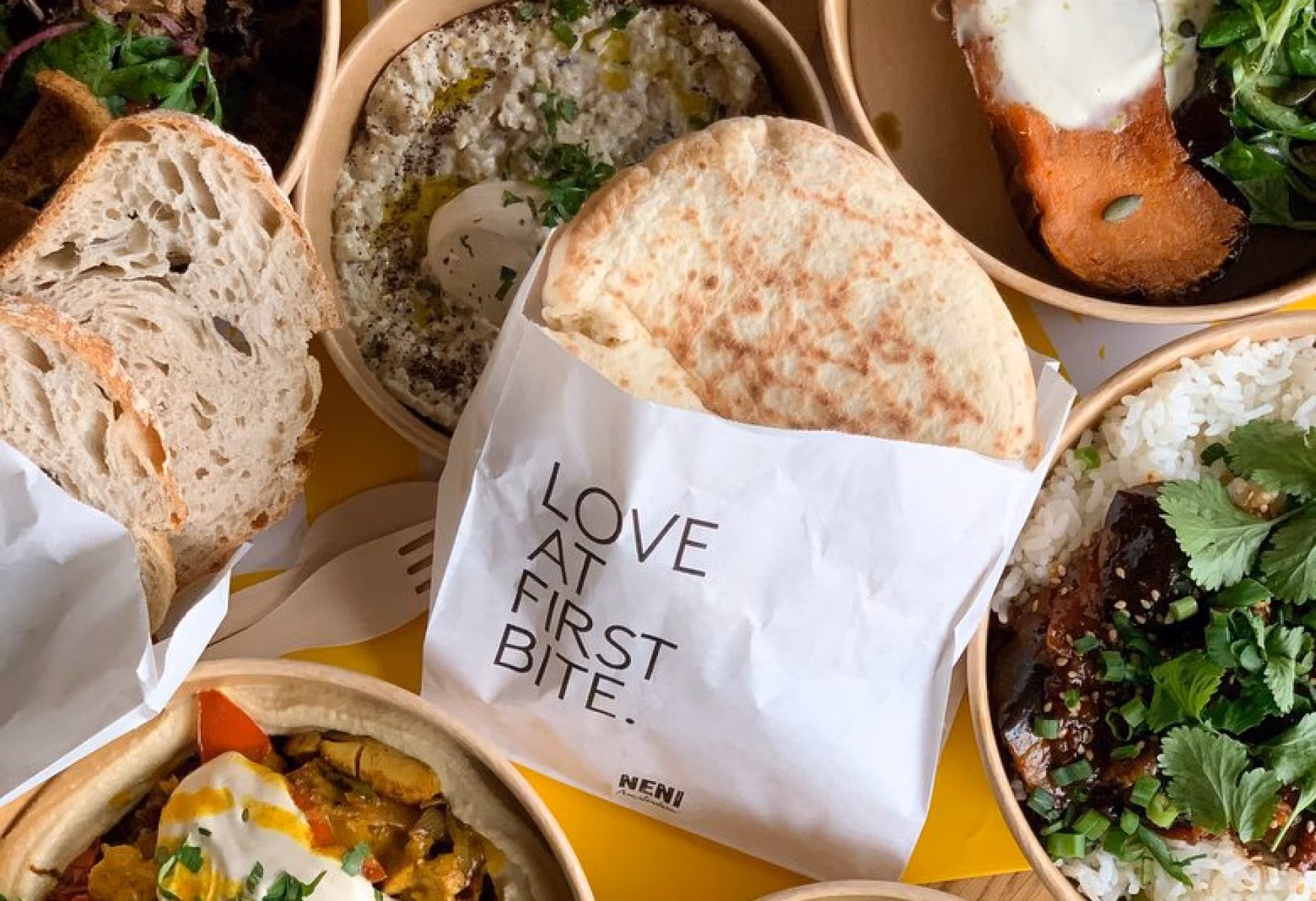 Takeaway spread with pita in a Love At First Bite paper bag surrounded by mezze bowls