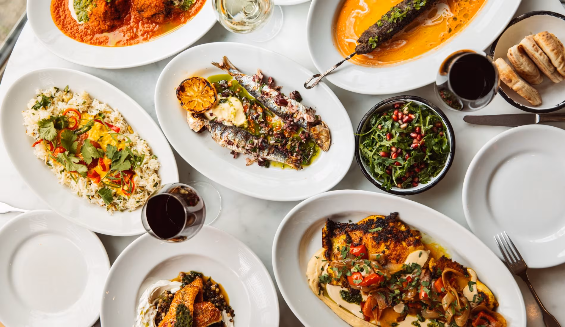 Overhead view of shared dishes with grilled aubergine, salads and wine