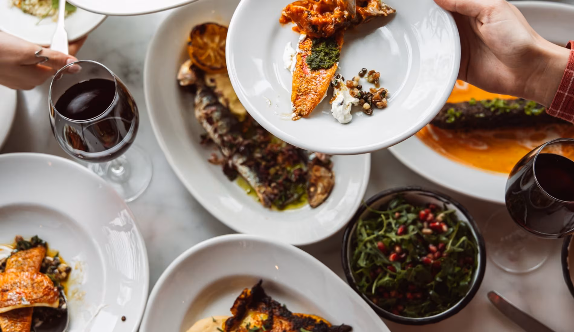 Guests reaching across a table of mezze and grilled dishes with red wine