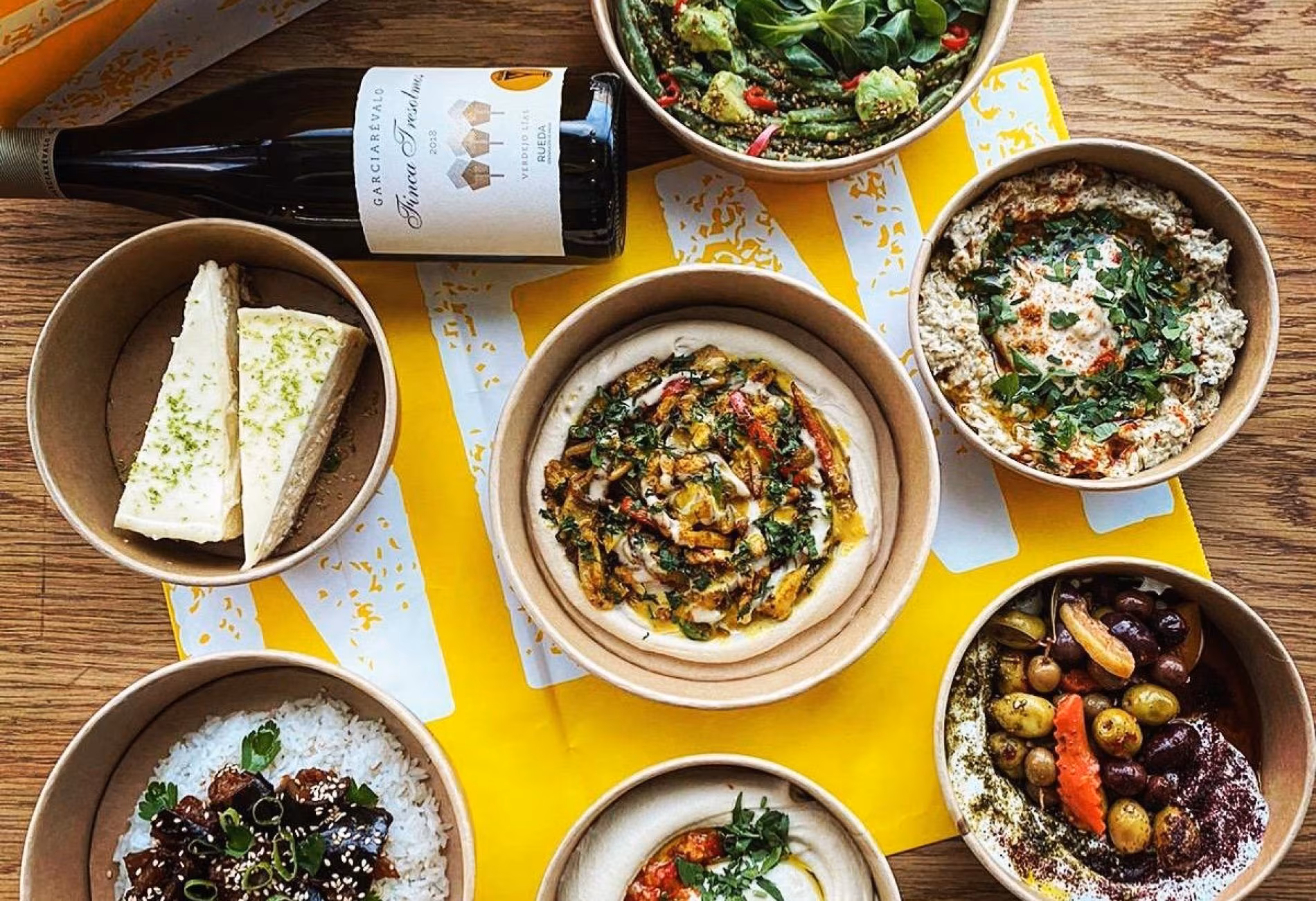 Salads, dips, bread and olives spread across a yellow table with a bottle of wine